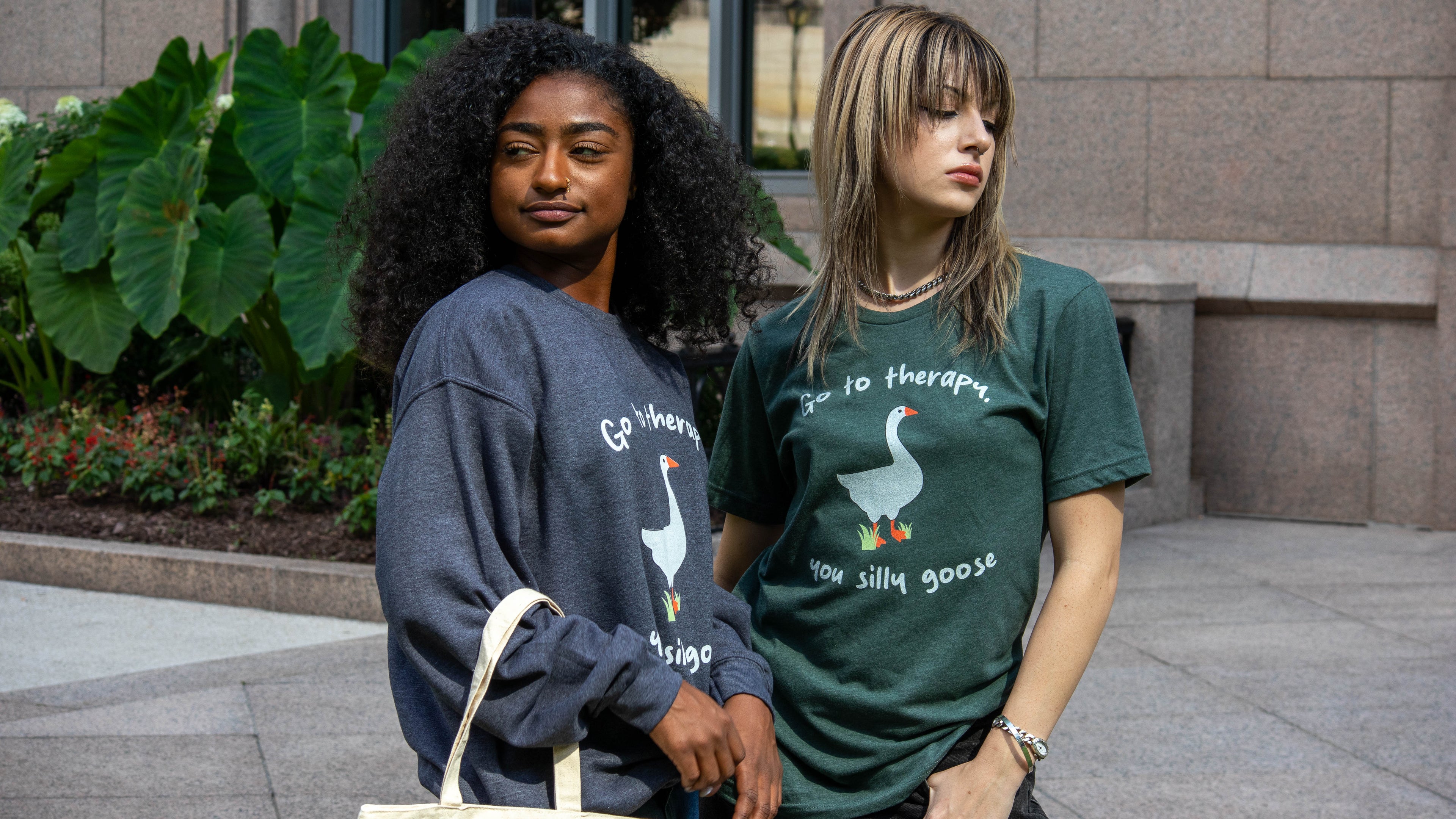 Two women wearing “Go to therapy, you silly goose” graphic tees pose confidently outdoors. One wears a dark gray long-sleeve, the other wears a green short-sleeve. Lush green plants and an office building are in the background.