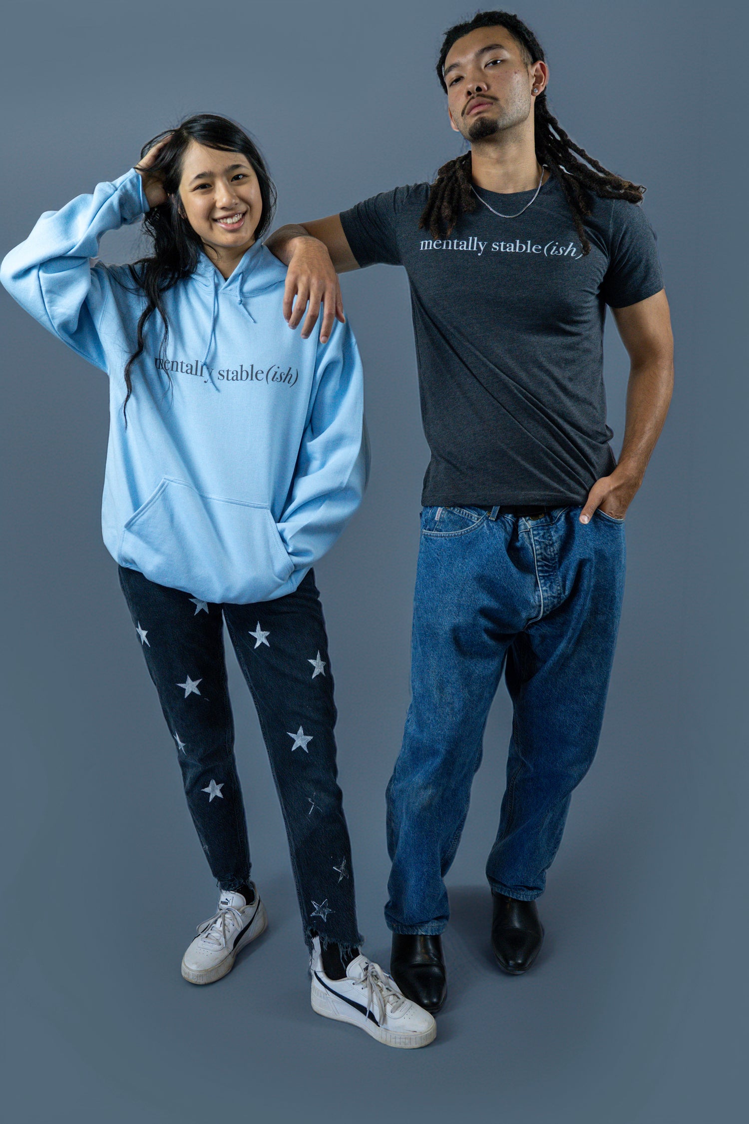 Two models wearing “mentally stable(ish)” apparel pose against a blue background—one in a light blue hoodie with star-patterned jeans, and the other in a dark charcoal t-shirt with relaxed blue jeans and black boots.