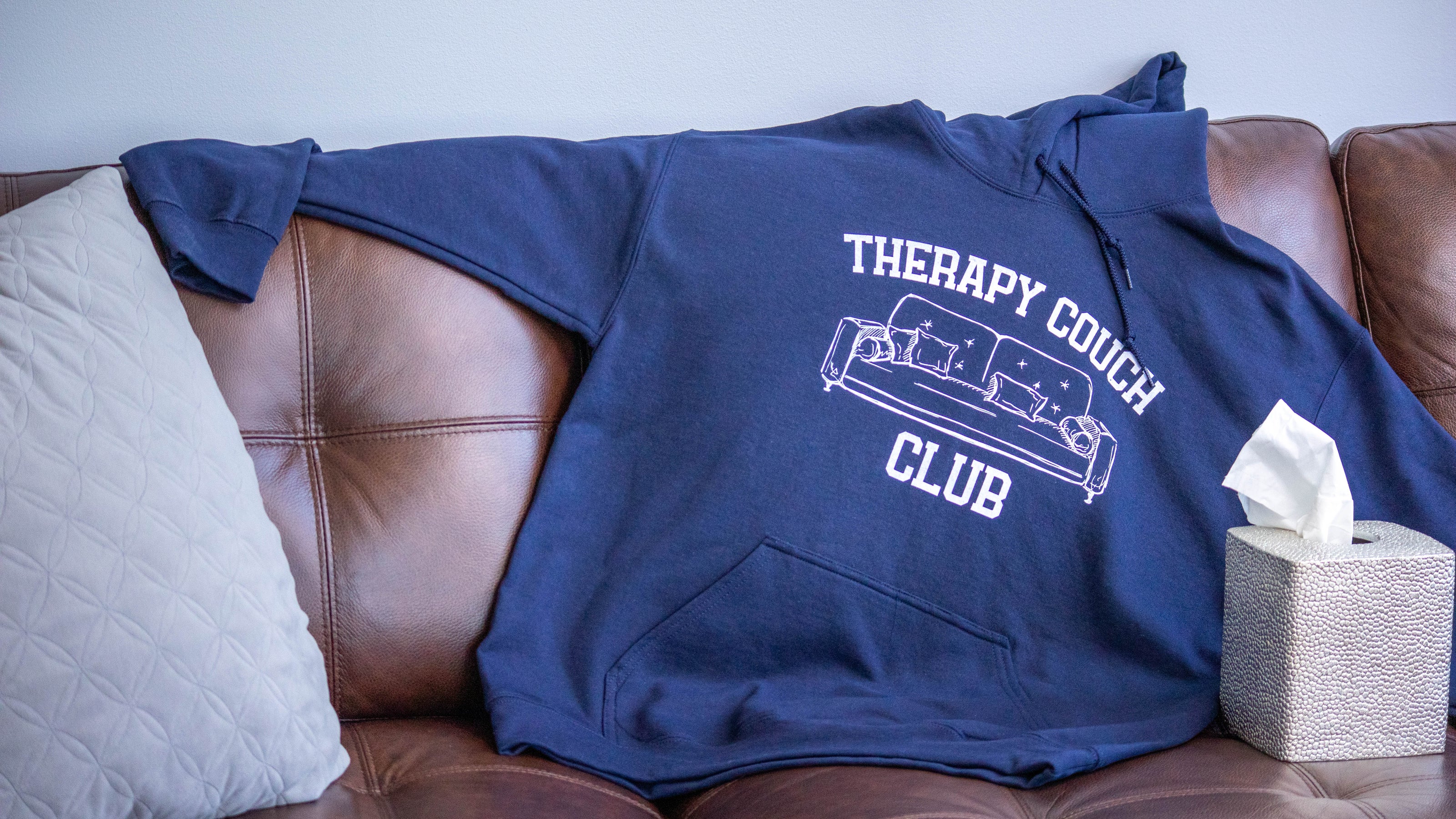 Navy blue “Therapy Couch Club” hoodie from Layered Feels draped on a leather couch with a tissue box and pillow, highlighting cozy mental health apparel that supports emotional wellness.