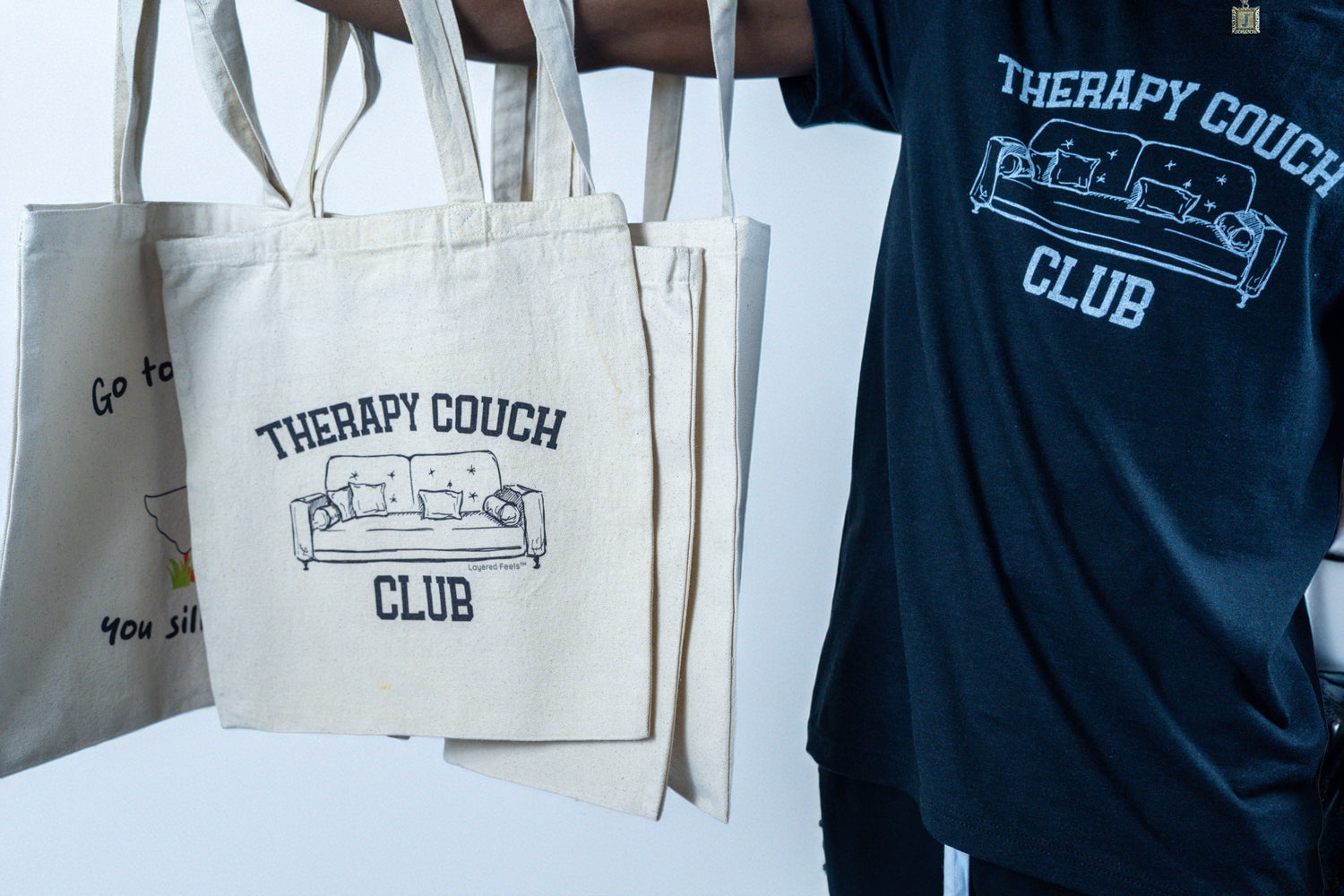 A close-up of a person holding multiple canvas tote bags featuring the “Therapy Couch Club” design, while wearing a matching black t-shirt with the same graphic.